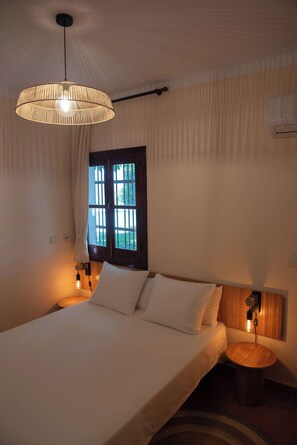 A bedroom with a bed, bedside lamps, a hanging light fixture, a window with curtains, and an air conditioning unit. - Finca Vinido is a charming, stylish holiday home close to Málaga and Marbella (Cartama)