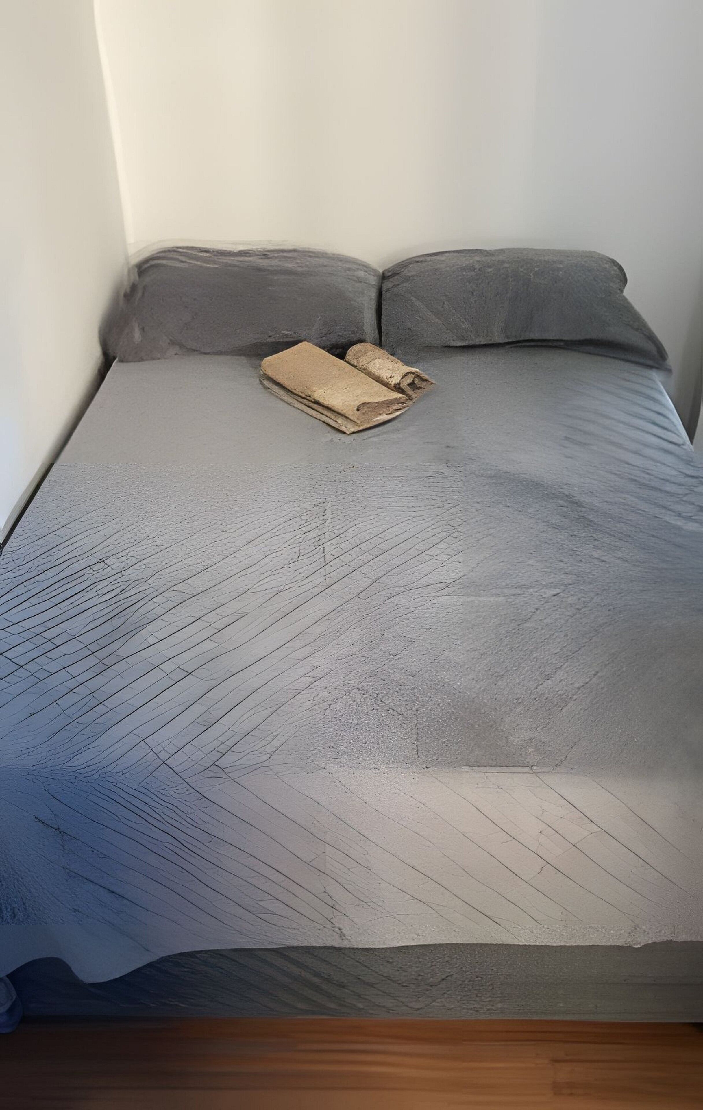 Ein Bett mit einer grau gemusterten Tagesdecke, zwei grauen Kissen und einer aufgerollten braunen Decke.