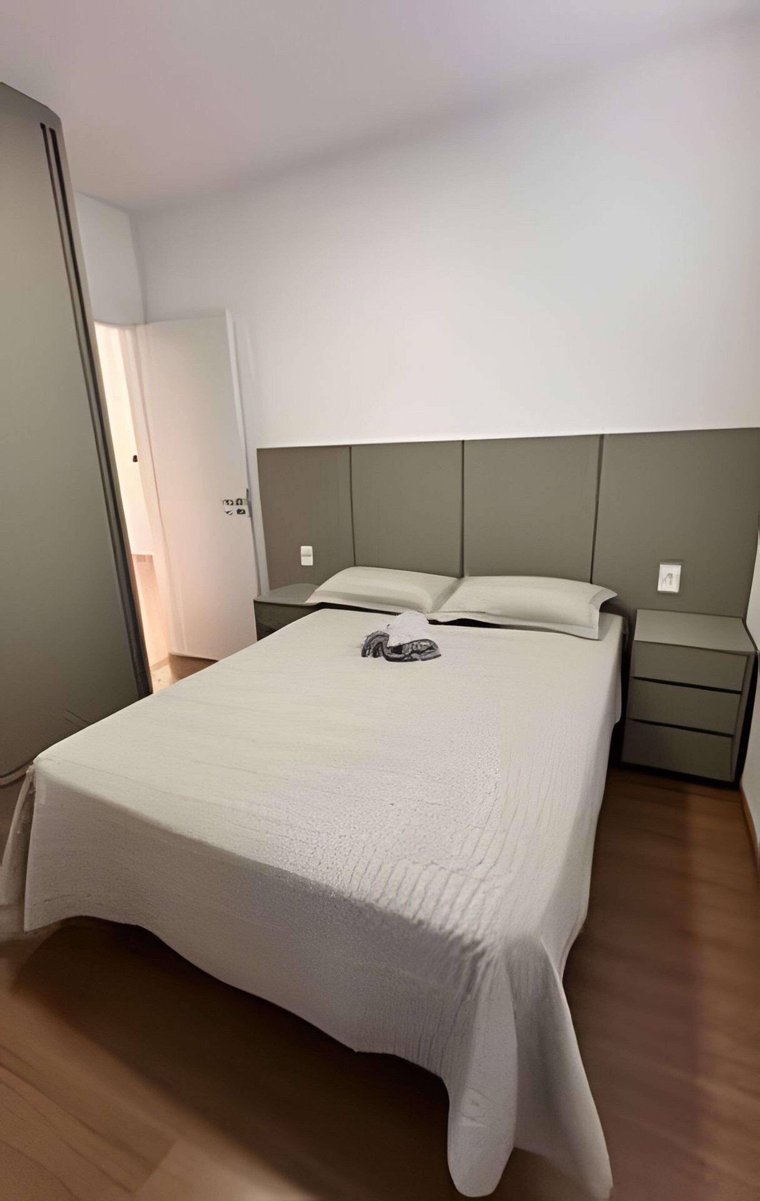 Una cama individual con una colcha blanca, dos almohadas, una mesita de noche y un pequeño mueble auxiliar.