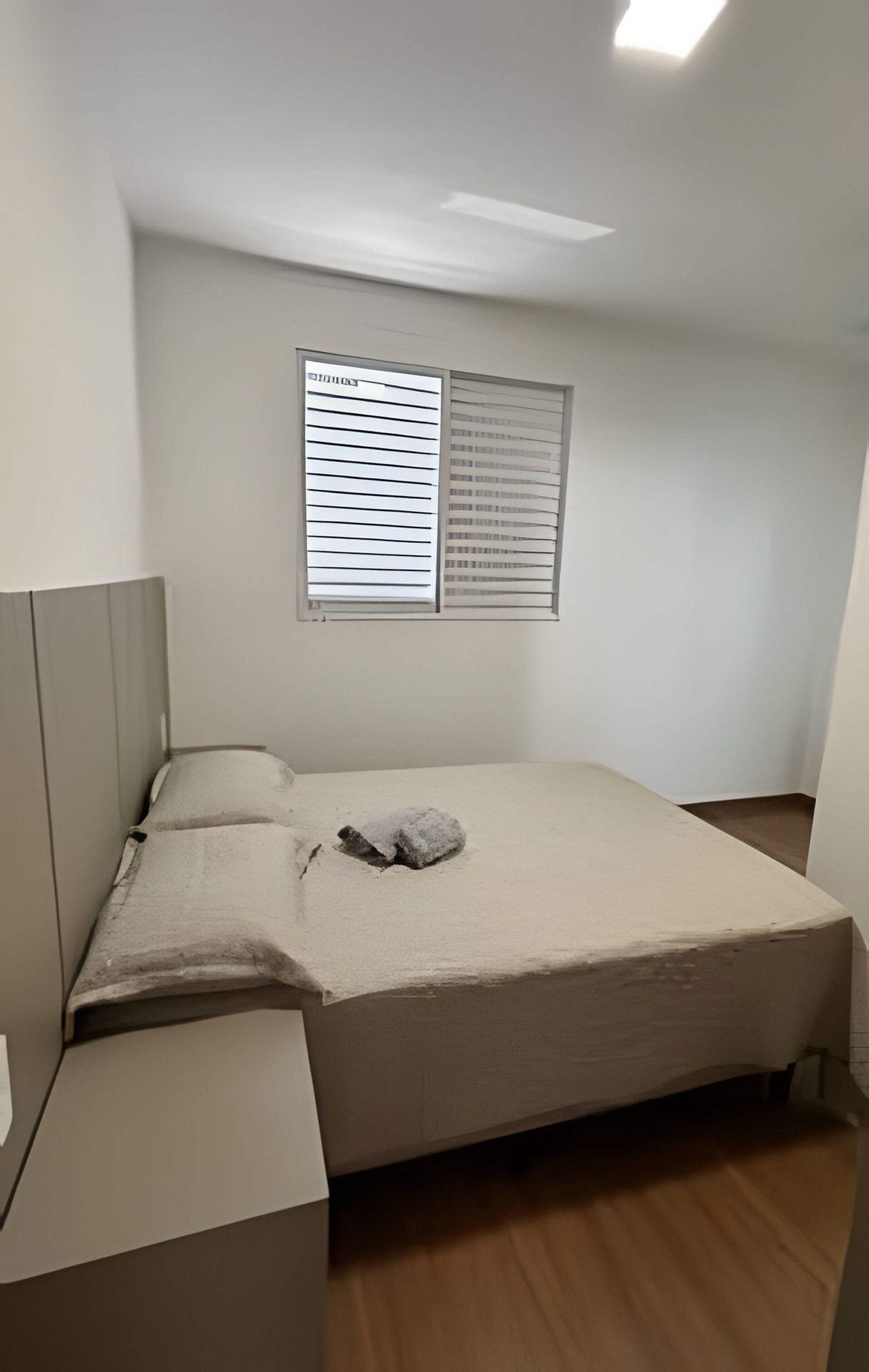 A small, single-bed room with a wooden floor, a bed with a beige mattress and pillows, a nightstand, and a window with blinds.