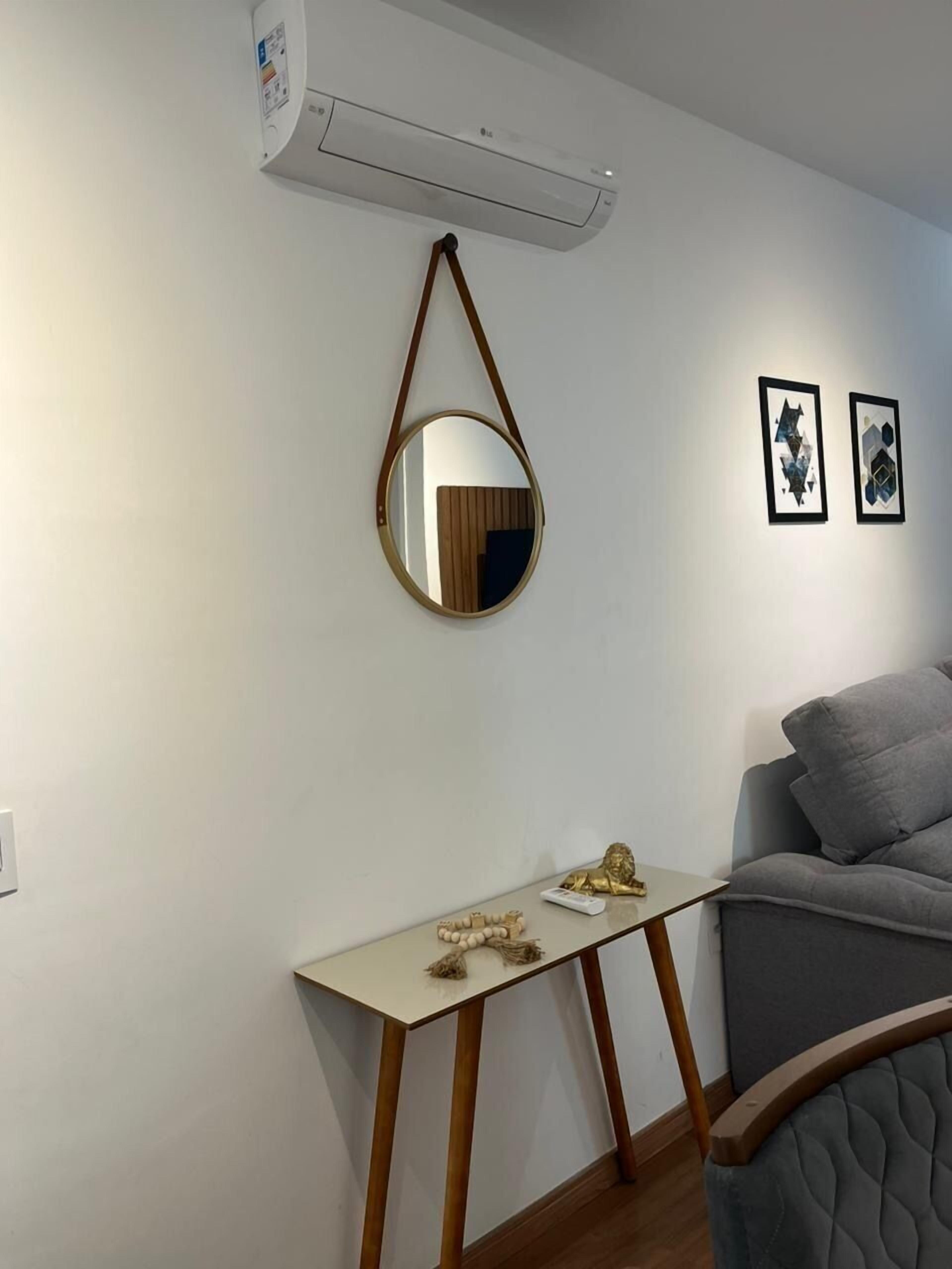 Una habitación con aire acondicionado blanco, un espejo redondo, una pequeña mesa con adornos y un sofá gris.