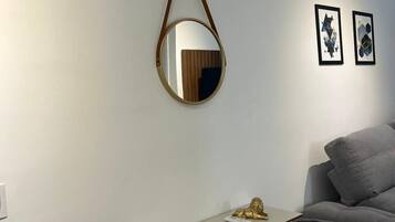 Una habitación con aire acondicionado blanco, un espejo redondo, una pequeña mesa con adornos y un sofá gris.