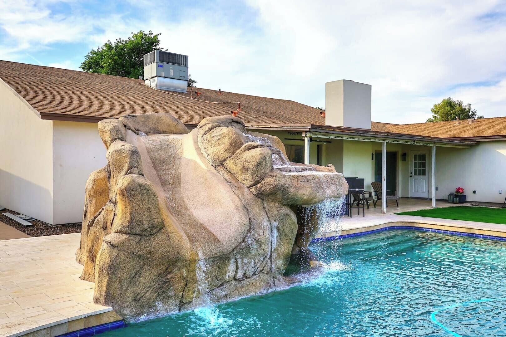 A swimming pool with a rock waterfall feature, a covered patio area, and a house with a tiled roof.