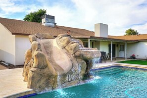 Une piscine agrémentée d’une cascade artificielle en roche, un patio couvert et une maison avec un toit en tuiles.