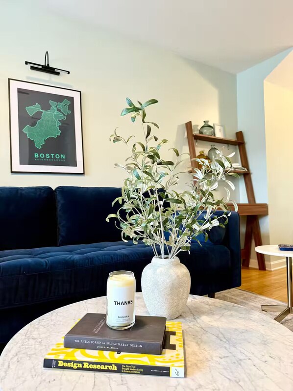 A living room with a dark blue sofa, a white vase with green foliage, a candle labeled "THANKS," and two books on a marble coffee table. - Boston Single House 2BR - Backyard + Walk to BCEC & Seaport (Boston)