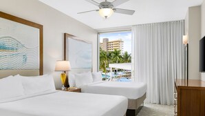 Quarto de hotel com duas camas, um ventilador de teto, uma janela grande com vista para palmeiras e uma piscina, e uma televisão do lado direito.