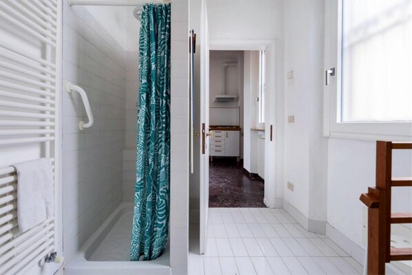 A bathroom with a shower, a towel rack, and a curtain. There is a hallway leading to a kitchen area.
