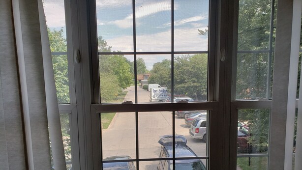Une vue à travers une fenêtre à carreaux, donnant sur un parking où se trouvent plusieurs voitures et un fourgon blanc avec des panneaux publicitaires bien visibles.