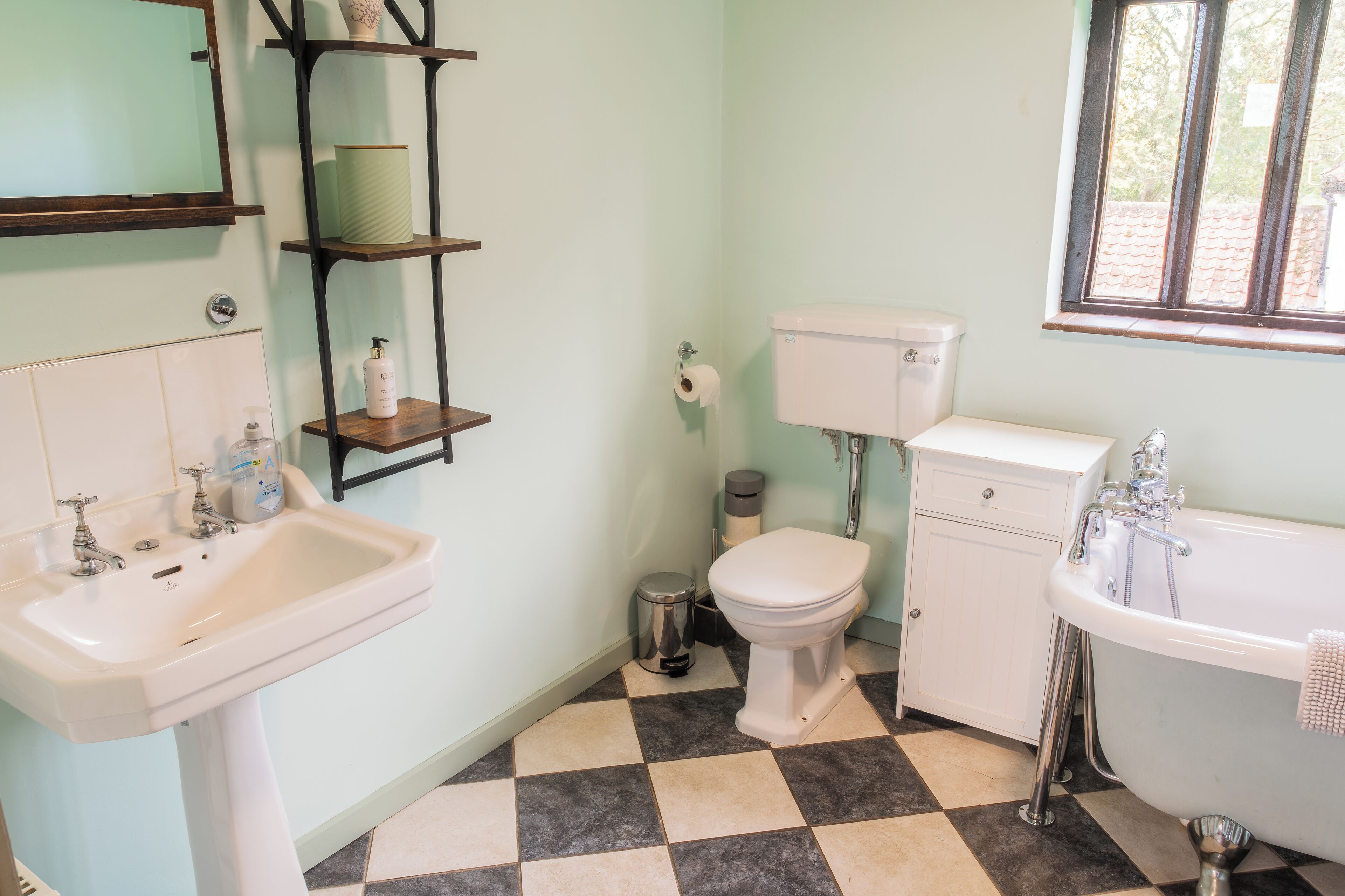 A bathroom with a sink, toilet, and bathtub. There is a shelf with toiletries and a trash can.