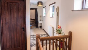 Un escalier en bois avec une main courante, une porte en bois sombre, un radiateur blanc, une petite table avec un vase de fleurs et une fenêtre avec une plante.