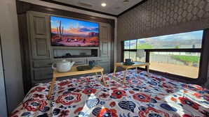A room with a large TV mounted on the wall, a bed with a floral-patterned bedspread, a small table with a teapot and cups, and a window with a view of a desert landscape. - Luxury Glamping Oasis: Superstition Mtns, Views & Nearby Lakes (Apache Junction)