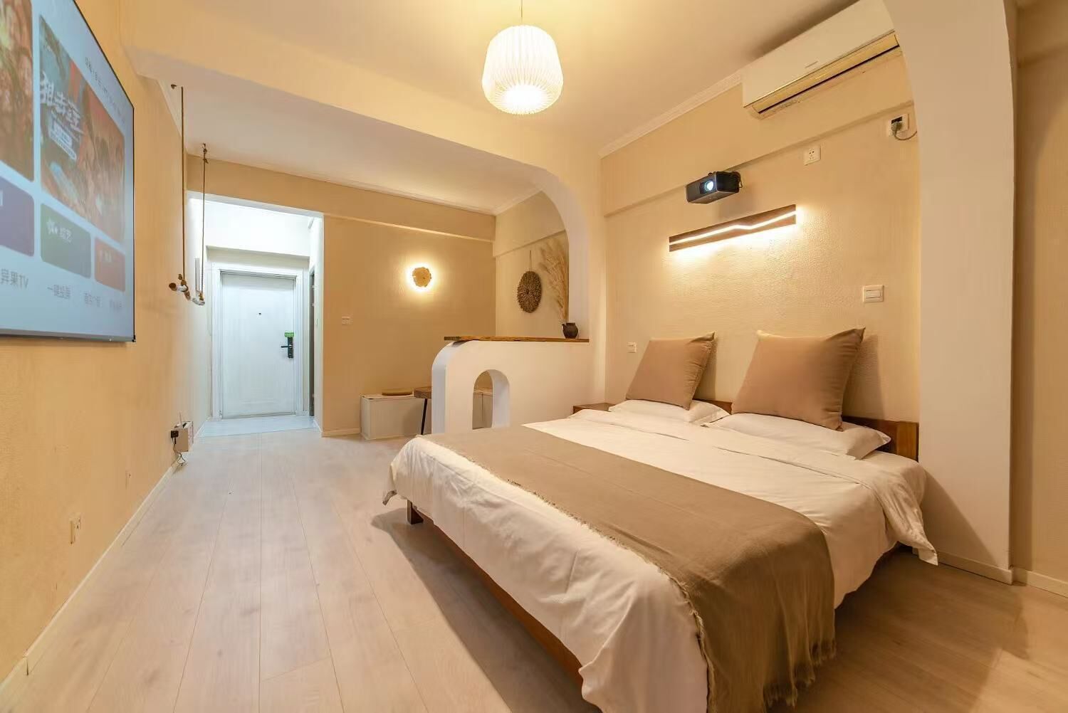 Habitación de hotel con una cama grande, un escritorio y un pequeño rincón de estar. Se ve una puerta que conduce a otra habitación y un proyector instalado en la pared.