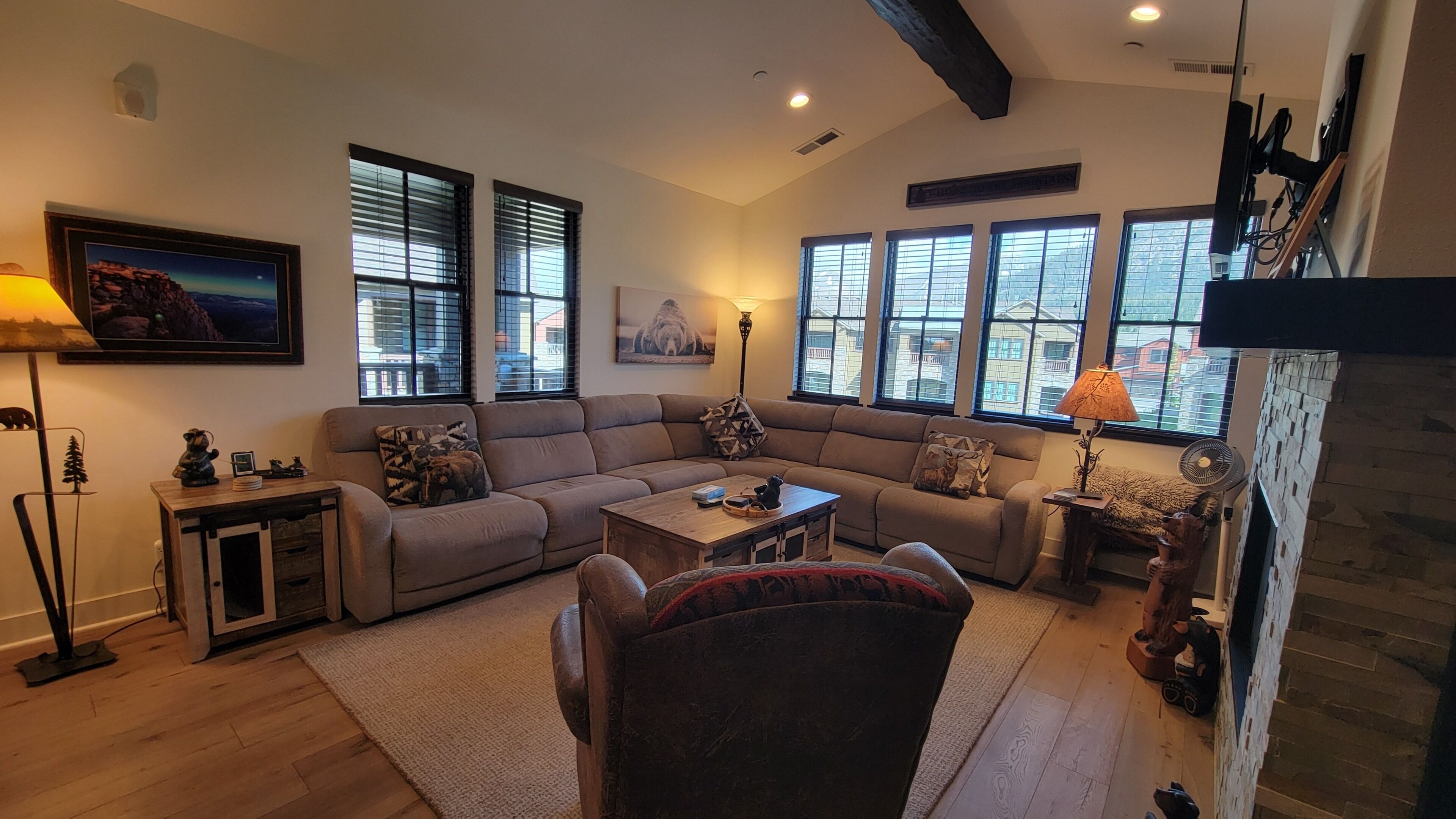 A living room with a sectional sofa, a coffee table, and a fireplace. There are large windows with blinds, a lamp, and a framed picture on the wall.
