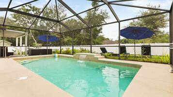 A covered swimming pool with two umbrellas, a lounge chair, and a small waterfall feature.