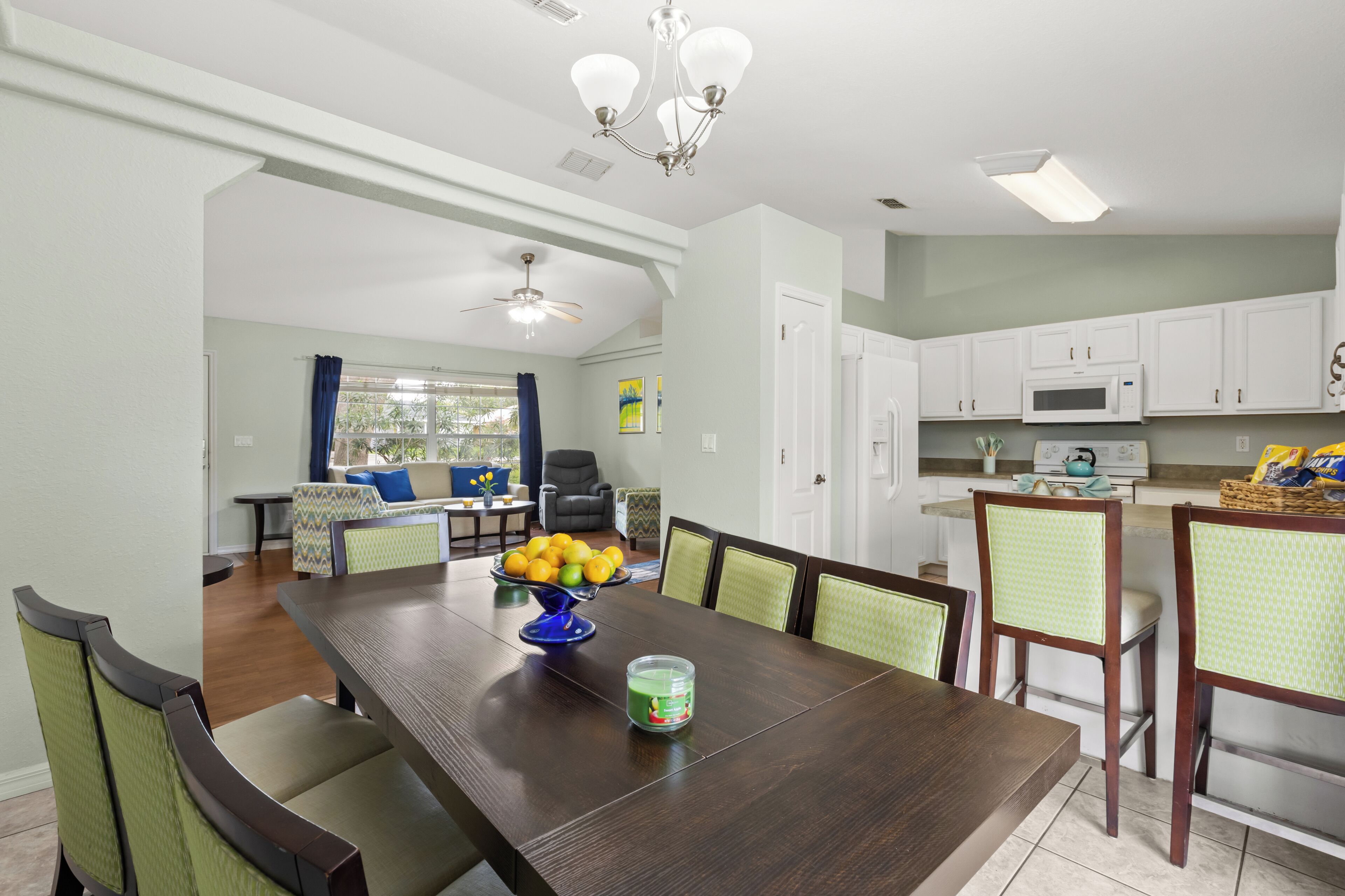 A dining area with a dark wooden table, green cushioned chairs, a blue bowl of fruit, and a green candle.