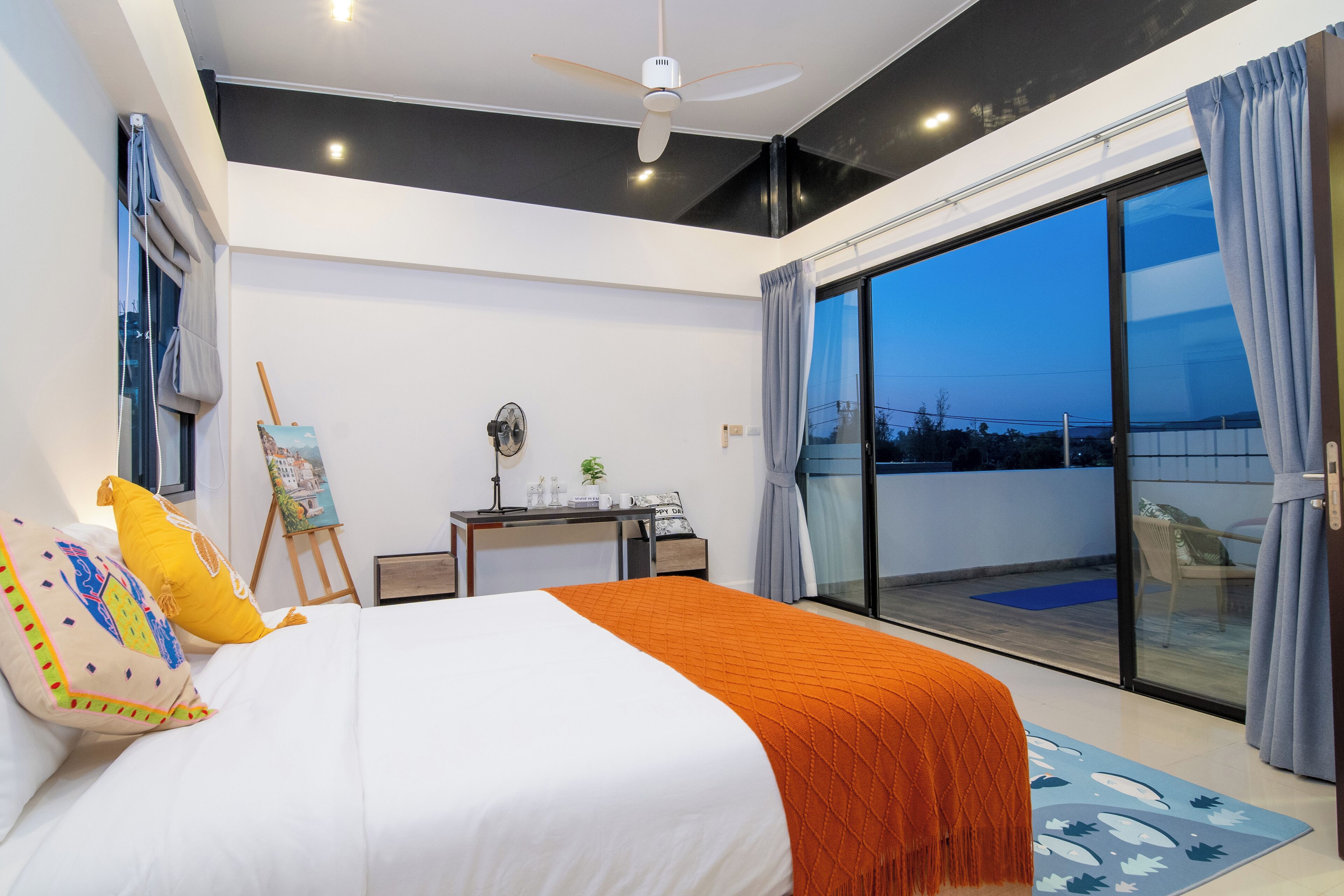 A modern bedroom with a large bed, a desk with a laptop, and a view of the outdoors through a sliding glass door.