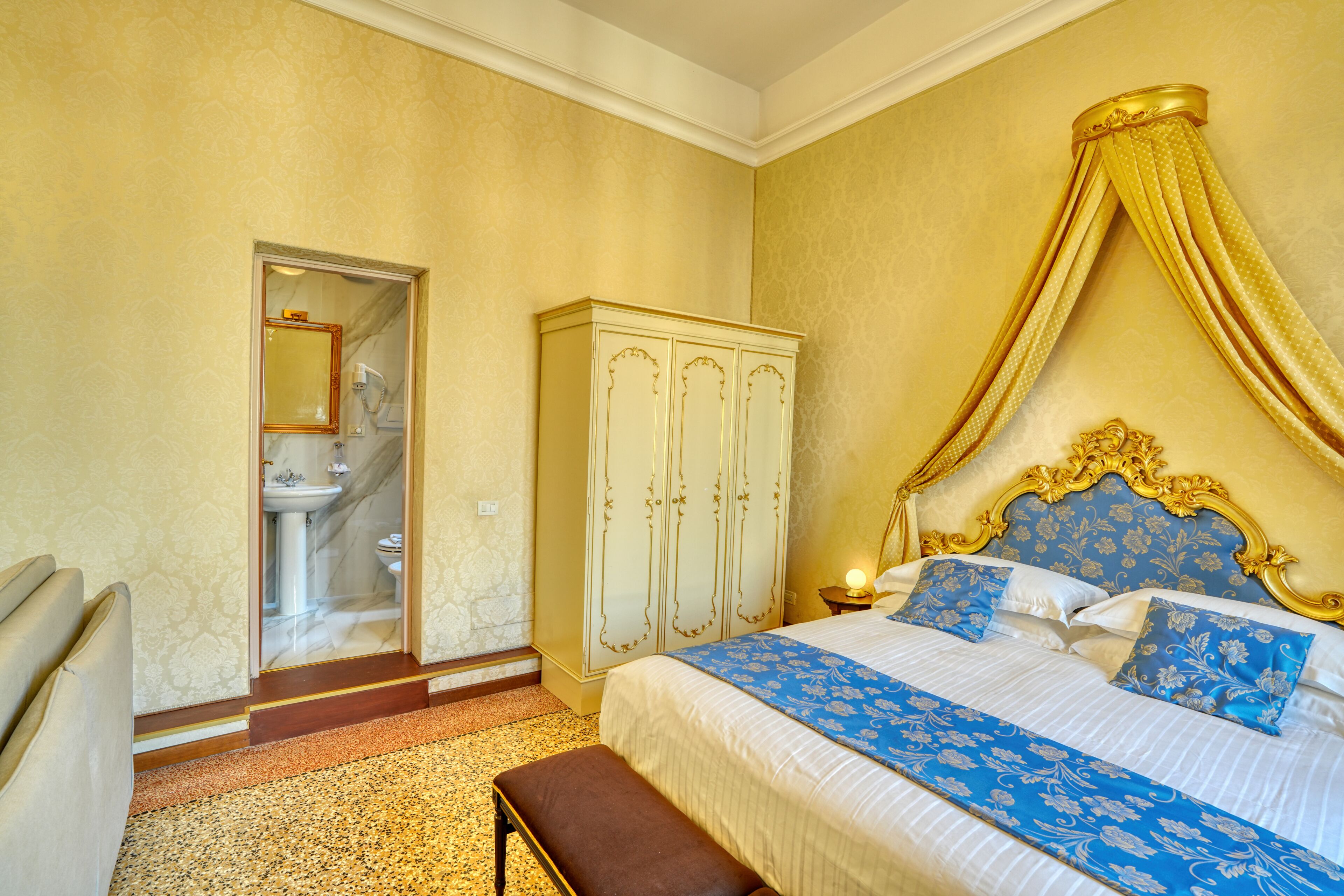 Ein Schlafzimmer mit Bett, Kleiderschrank, einer Bank und einem durchgehenden Türspalt sichtbaren Badezimmer.