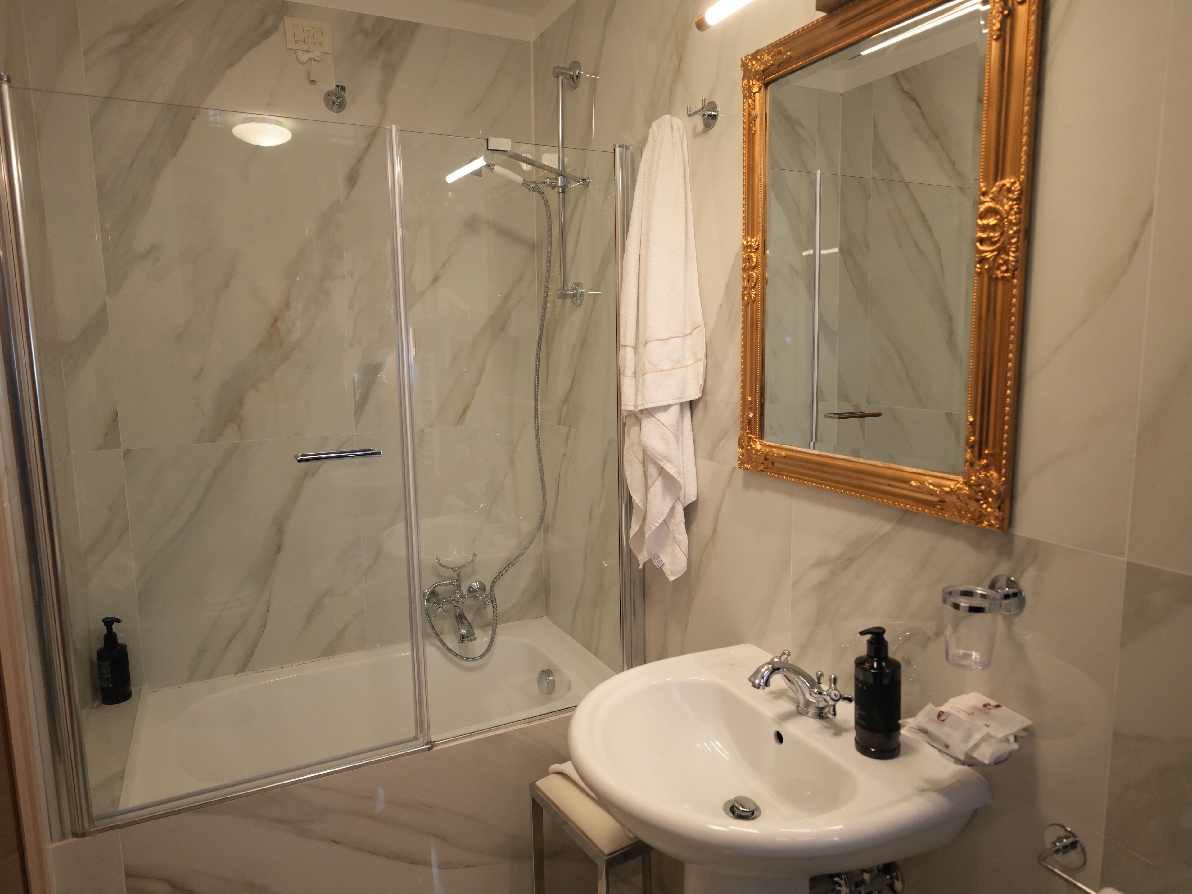 Une salle de bain dotée d’une douche à parois de verre, d’un lavabo blanc avec un miroir et de murs en marbre.