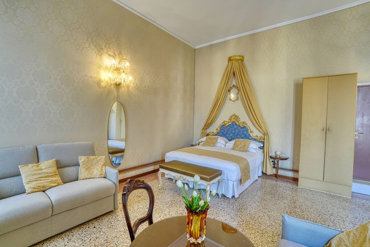A hotel room with a bed, a sofa, a dining table with a vase, a mirror, and a wardrobe.