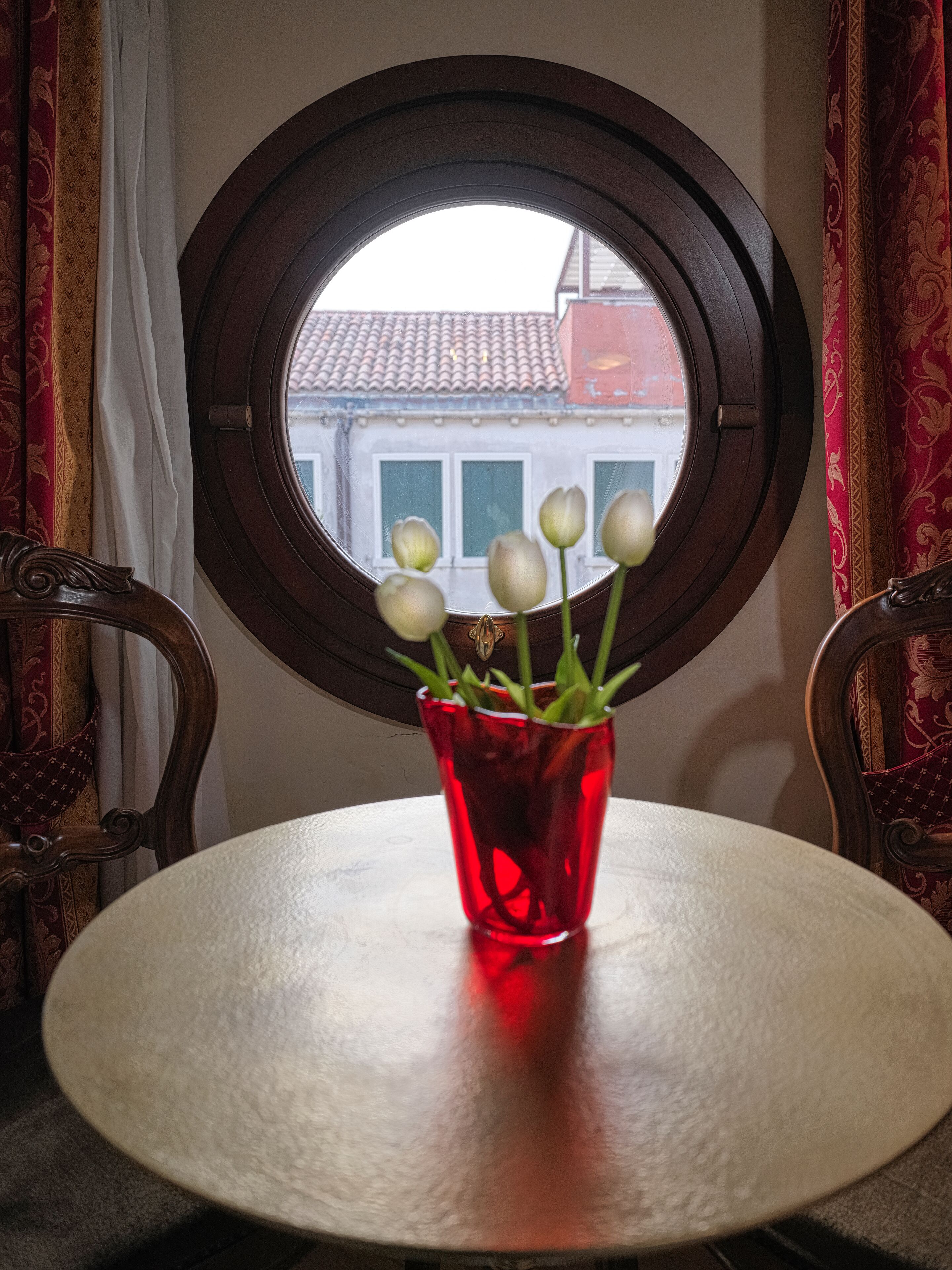 Uma mesa redonda com um vaso de vidro vermelho com flores brancas, colocada em frente a uma janela com moldura marrom.