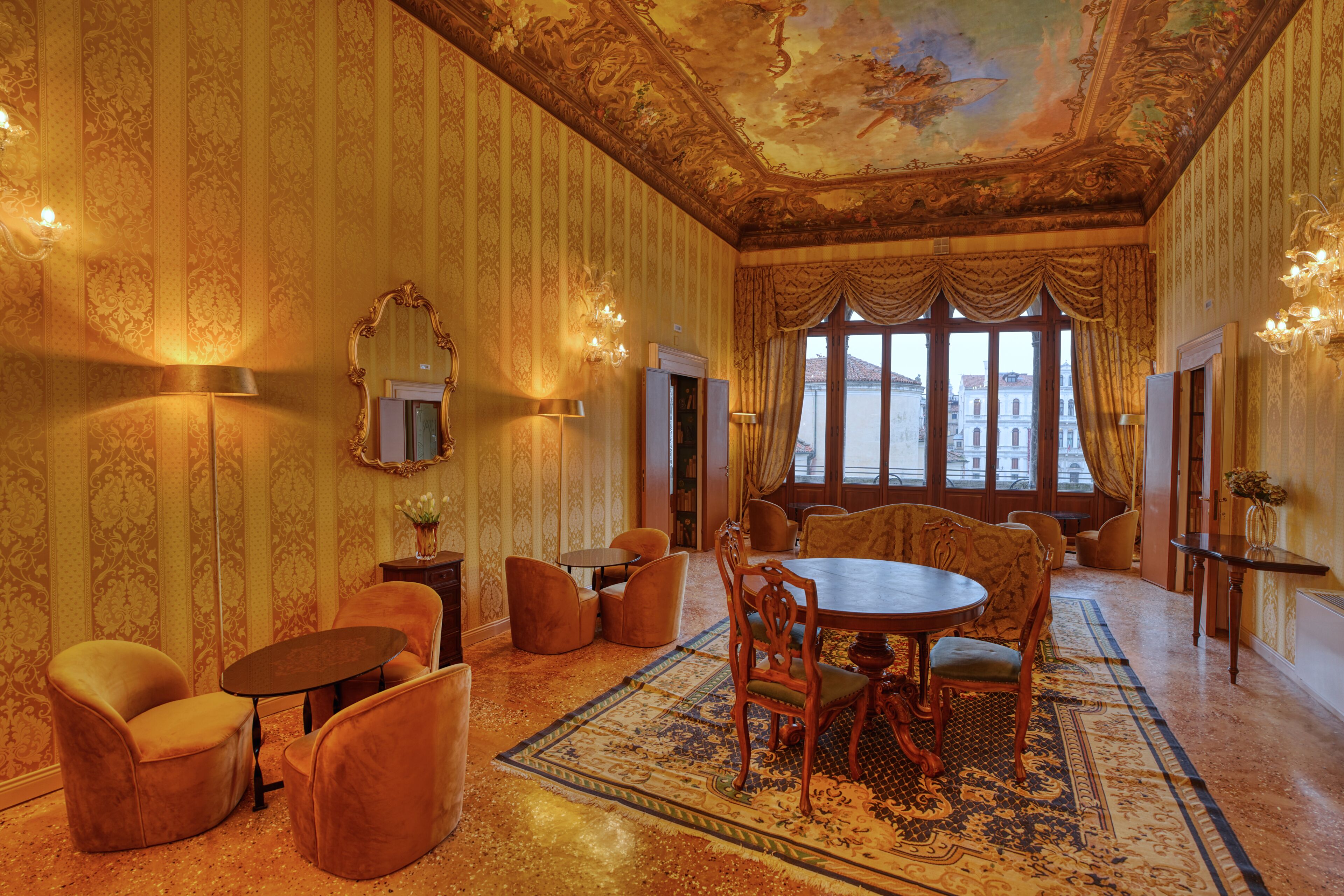 A room with a large window, ornate ceiling, and a dining area with chairs and a table.