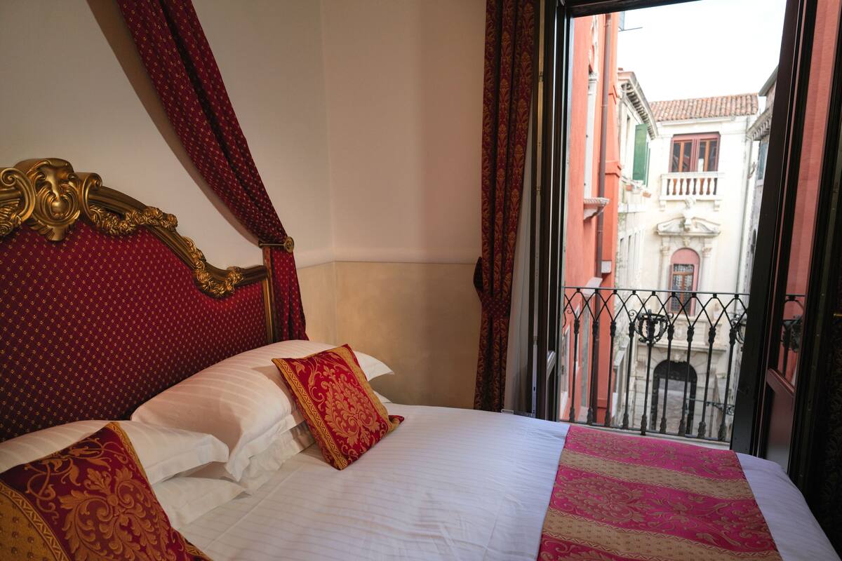 A hotel room with a large bed, a balcony with a wrought iron railing, and a view of buildings outside.