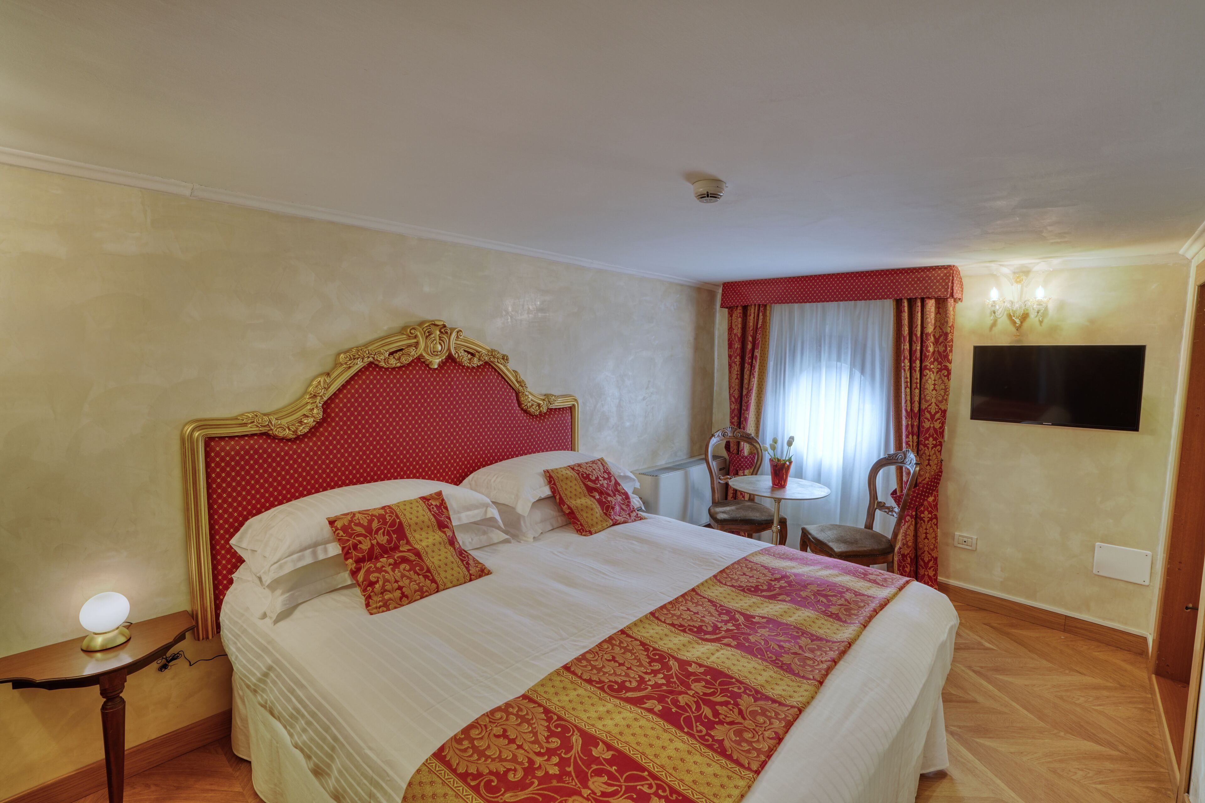 A hotel room with a large bed, a bedside table with a lamp, a chair, a small table, and a TV mounted on the wall.