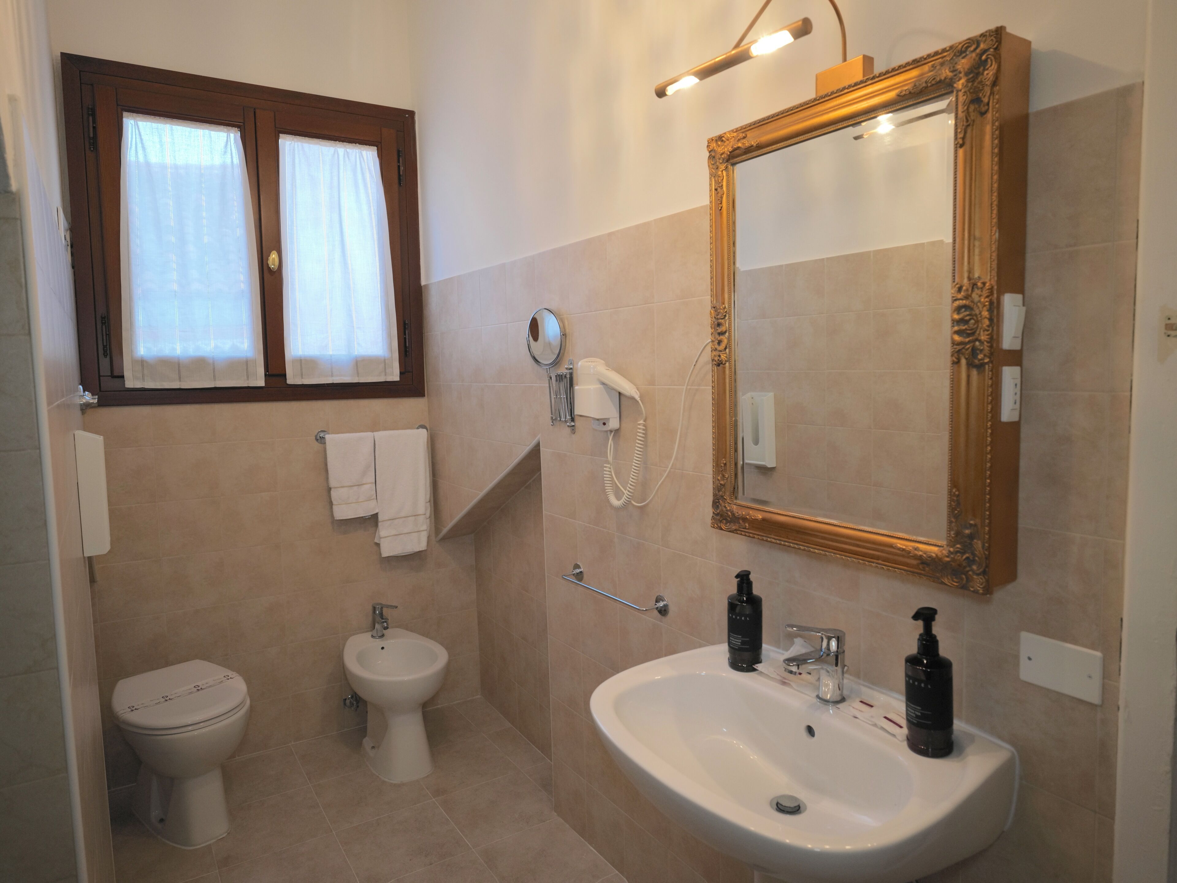 Une salle de bain équipée de toilettes, d’un bidet, d’un lavabo, d’un miroir et d’un sèche-cheveux fixé au mur.