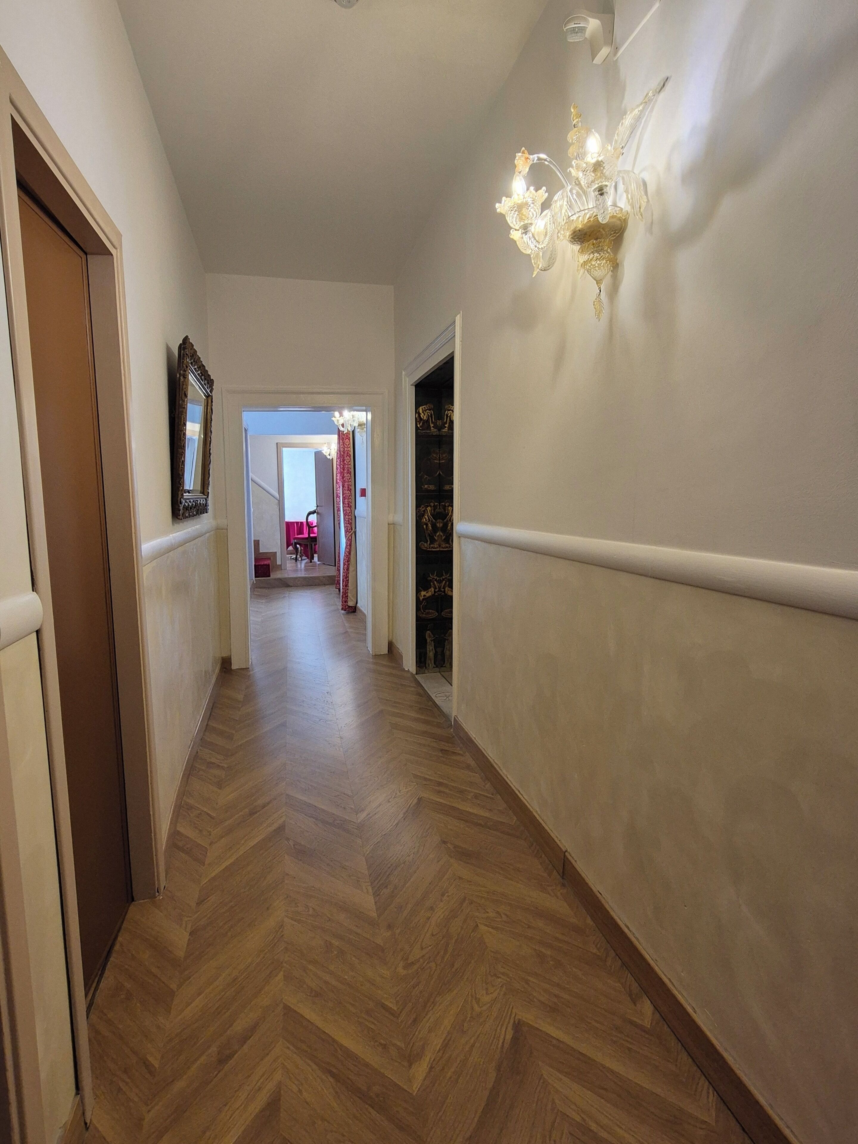 A hallway with a wooden herringbone floor, a wall-mounted light fixture, and a door on the left.