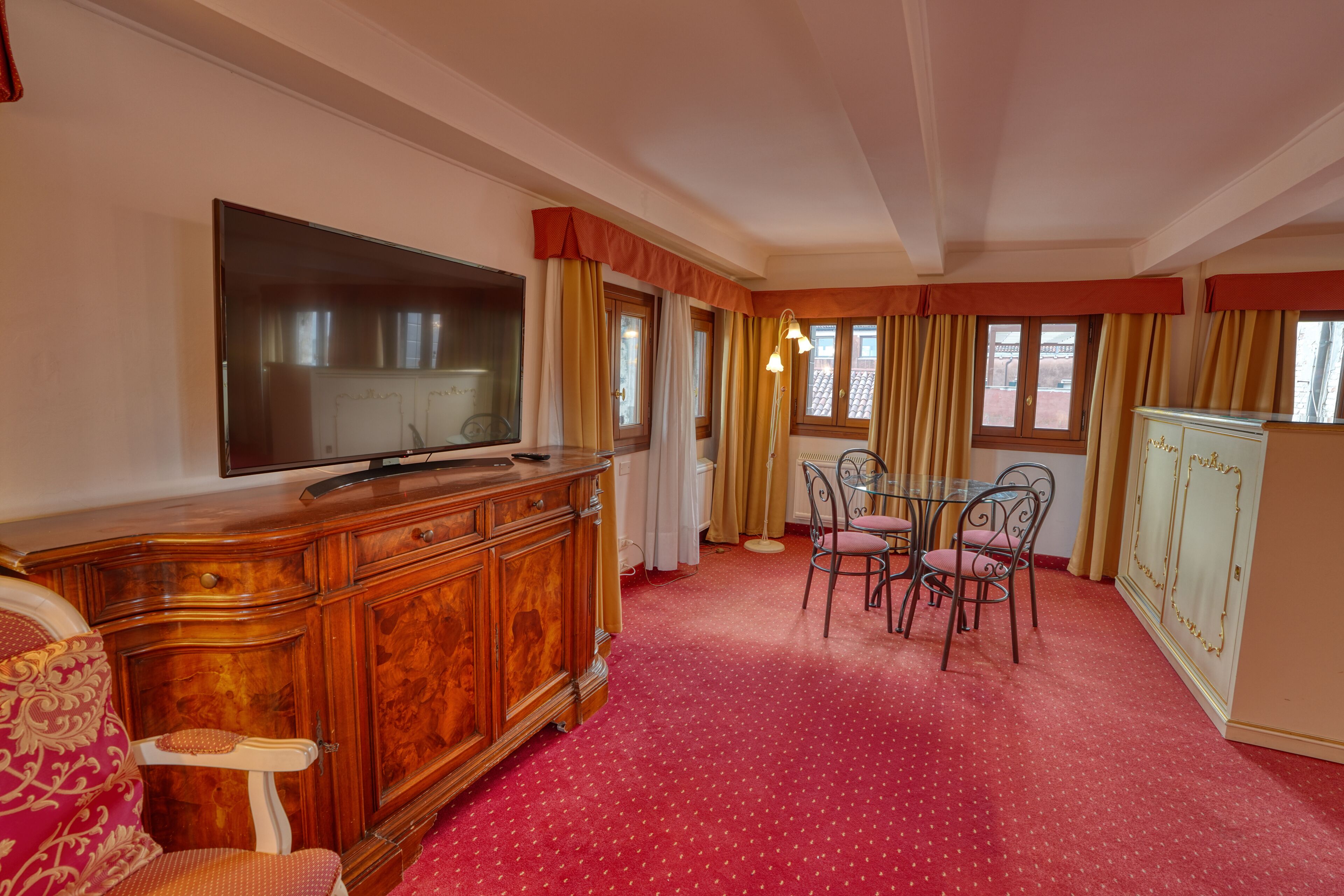 A room with a television, wooden cabinet, dining table with chairs, and a patterned carpet.