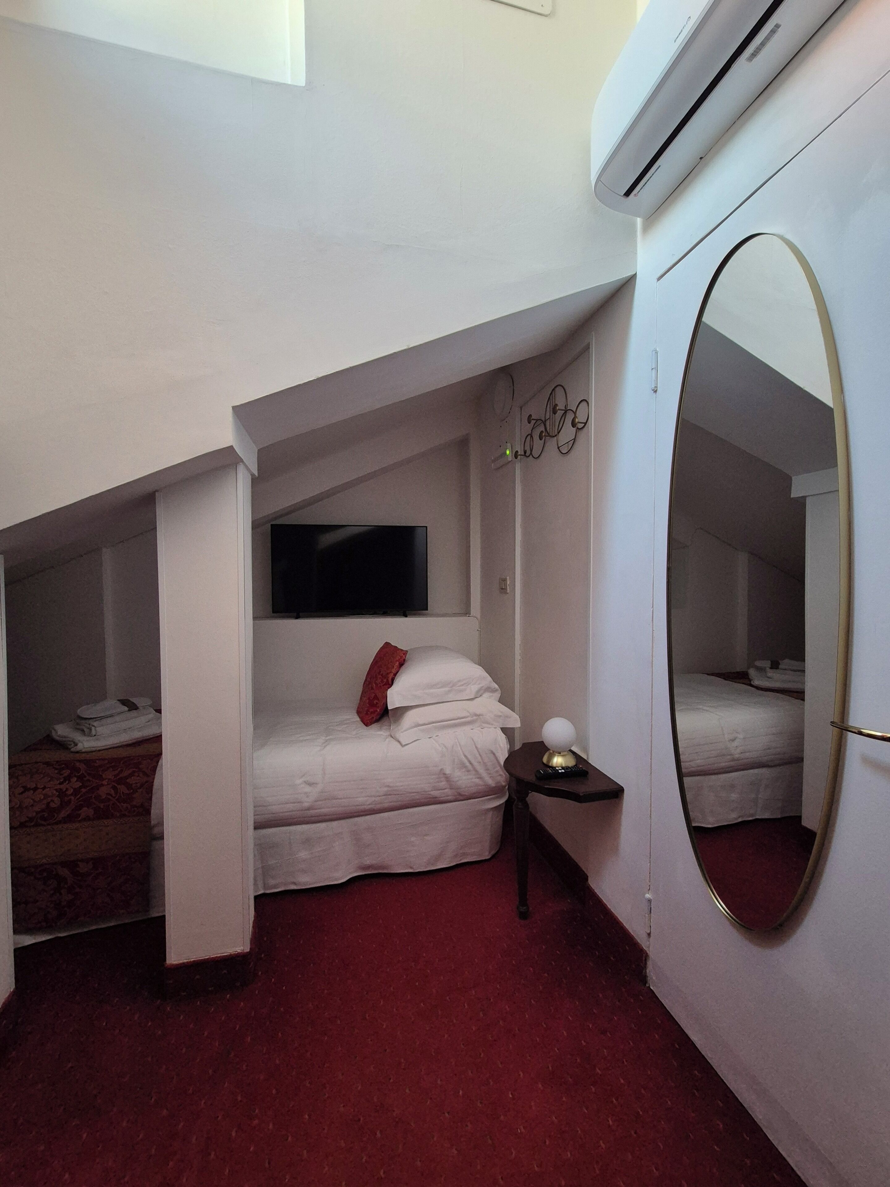 A small, compact room with a bed, a bedside table, a mirror, and a wall-mounted TV.