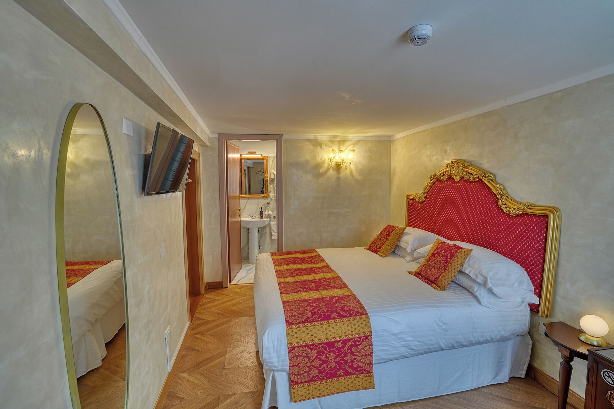 Une chambre d’hôtel avec un grand lit, un miroir, une table de chevet avec une lampe, et une salle de bain visible à travers une porte ouverte.