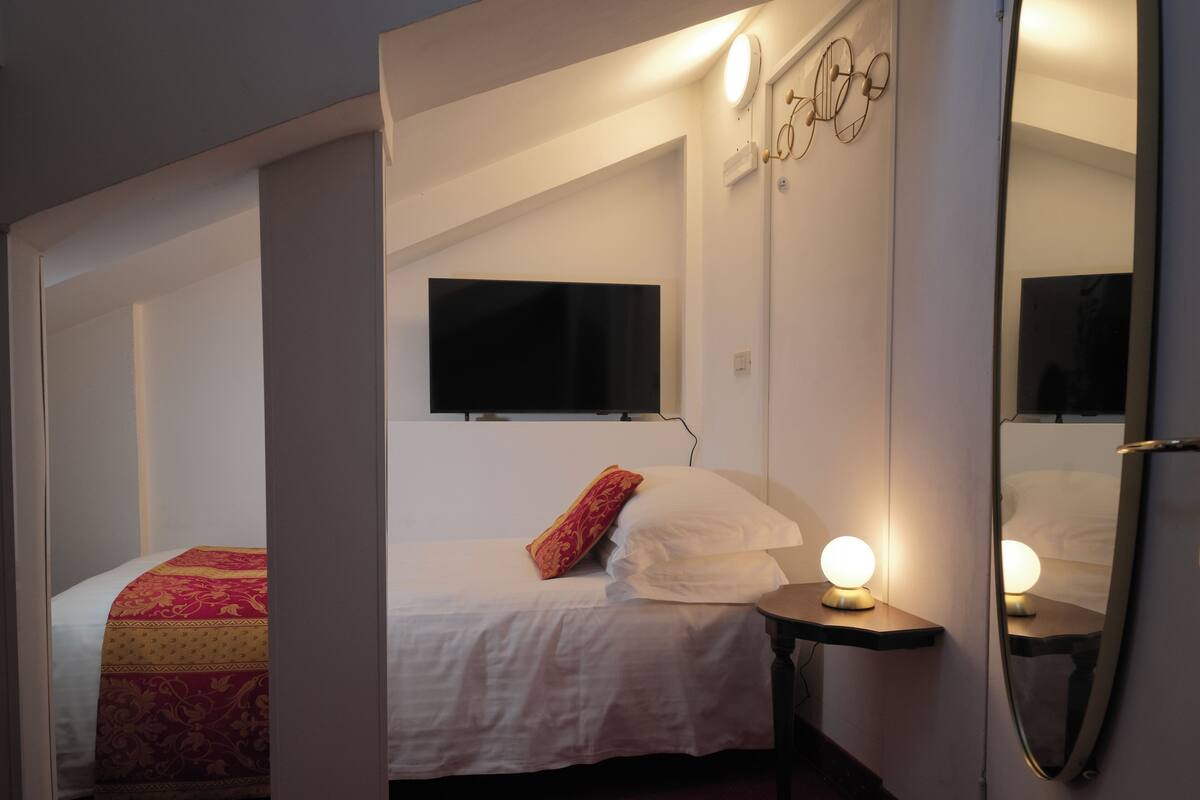 A bedroom with a bed, a bedside table with a lamp, a flat-screen TV mounted on the wall, and a mirror on the wall.