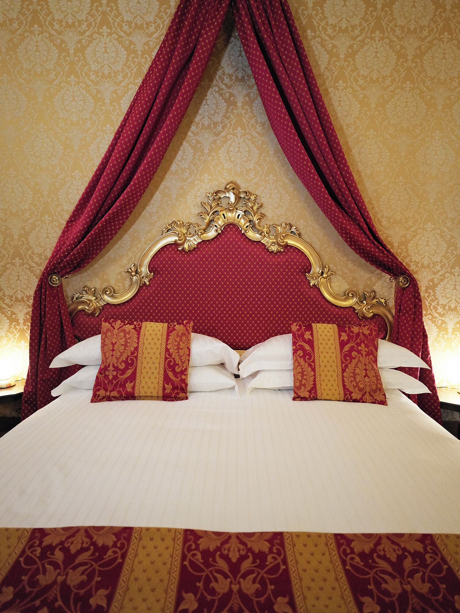 A bed with a red canopy and ornate gold headboard, white linens, and patterned pillows.