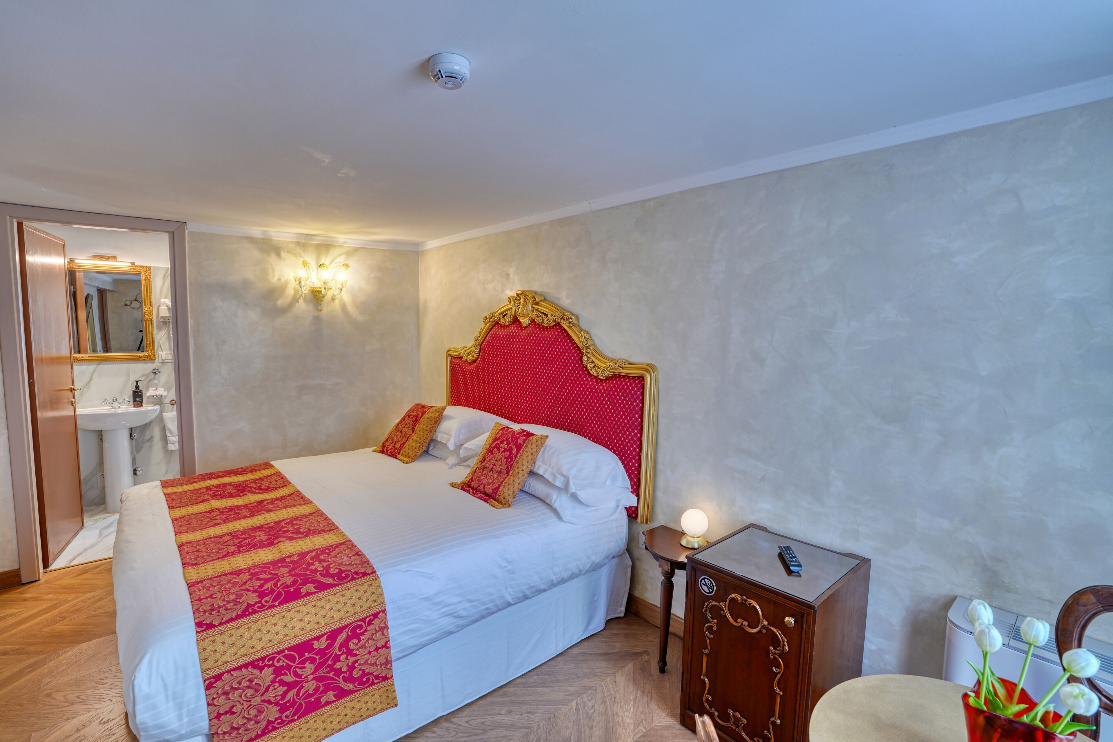 A hotel room with a bed, bedside table, lamp, and a vase with flowers.