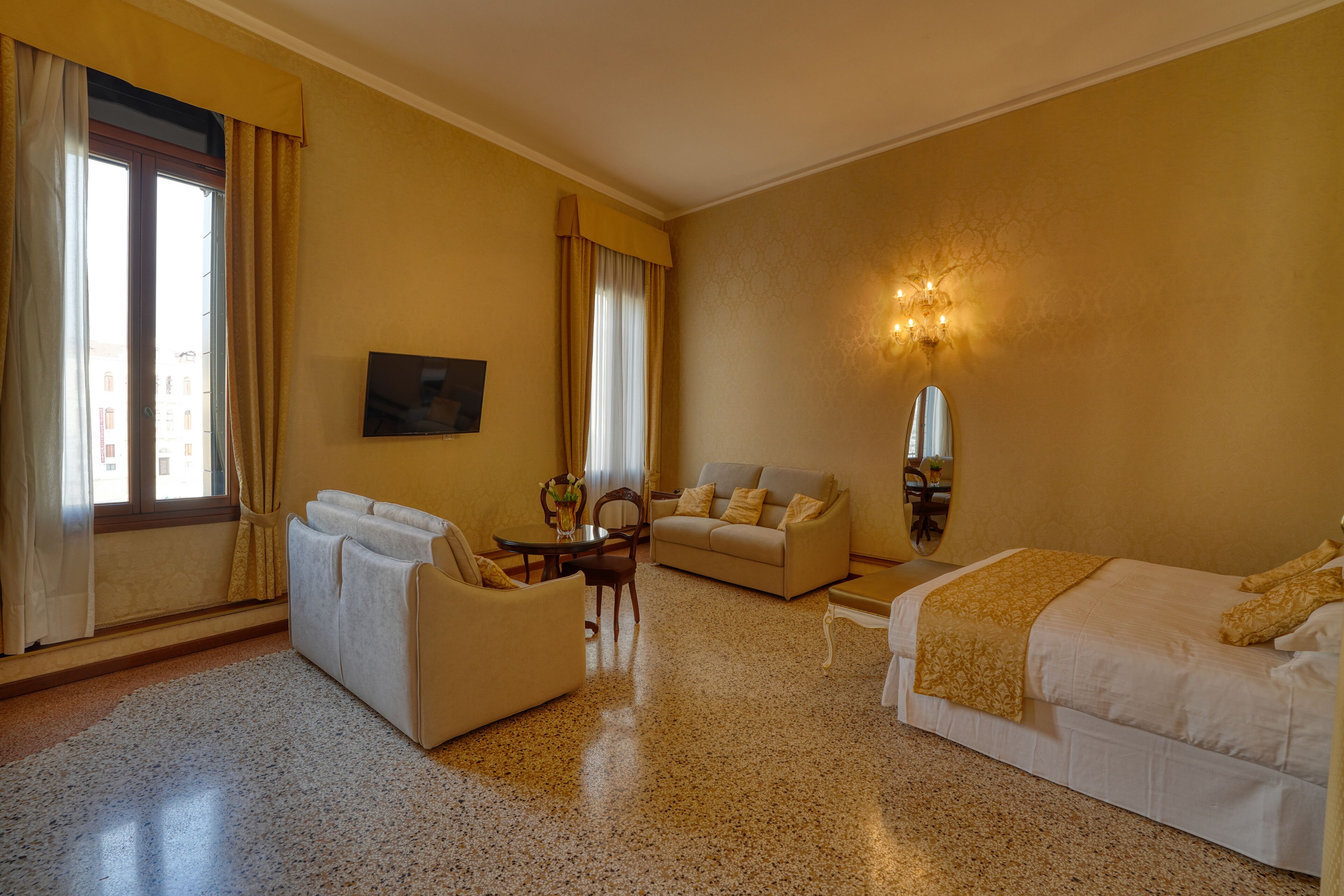 A hotel room with a bed, two sofas, a small table, a chair, a TV, and a window with curtains.