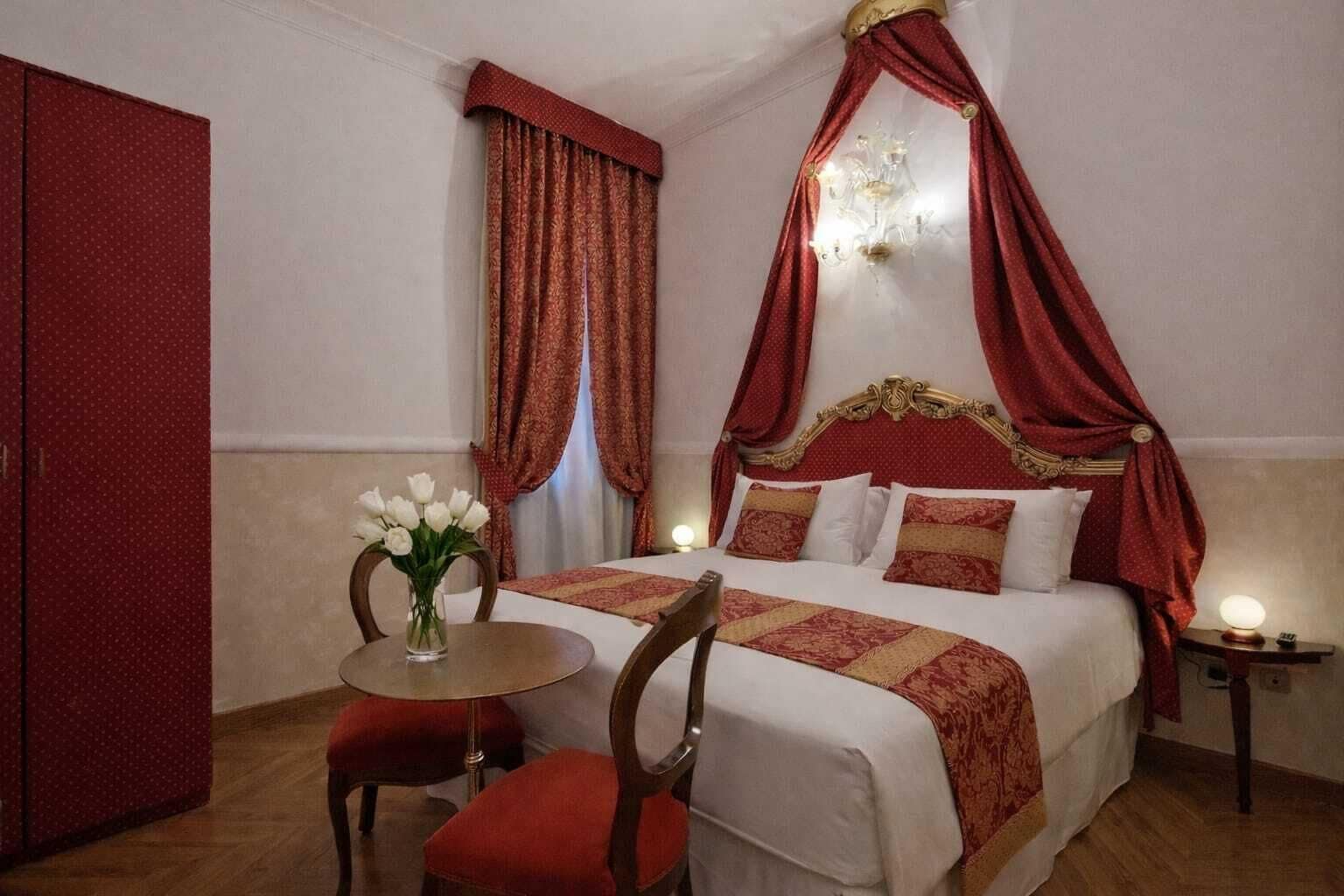 Une chambre d’hôtel avec un lit à baldaquin, un vase de fleurs, une table avec une chaise et une table de chevet avec une lampe.