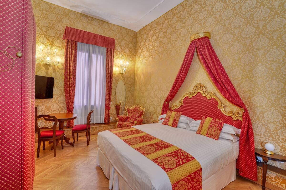 A room with a four-poster bed, a round wooden table with chairs, a television, and a chandelier.