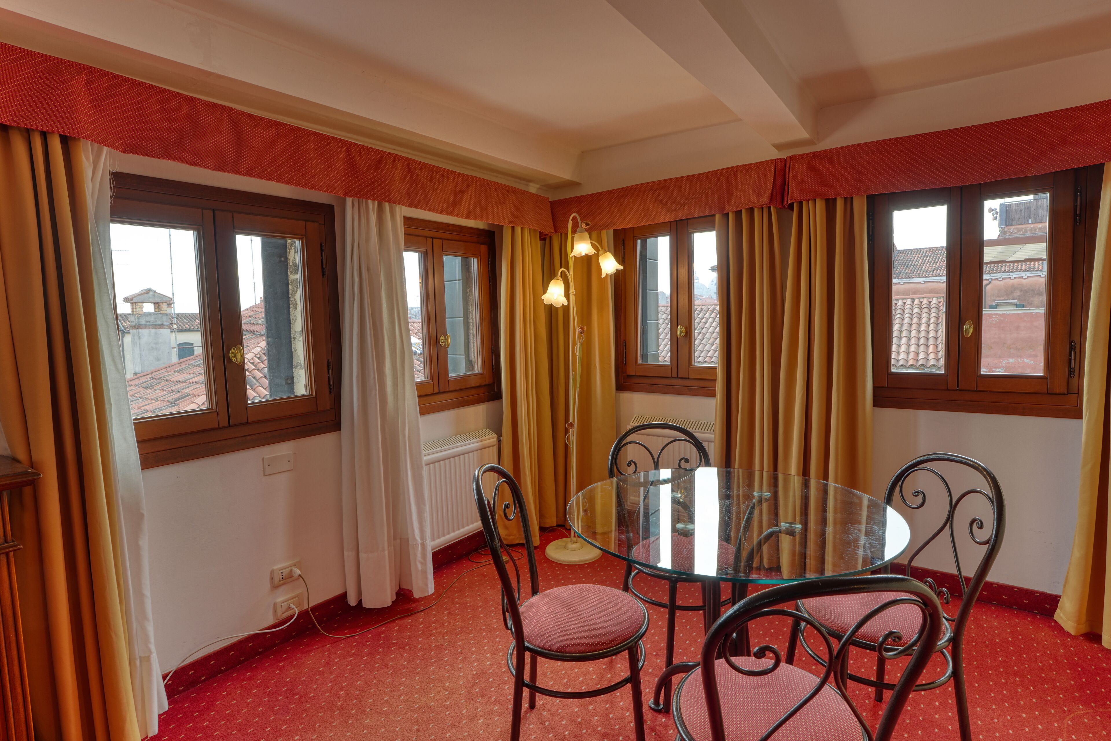 A dining area with a glass table and metal chairs, a radiator, and windows with curtains.