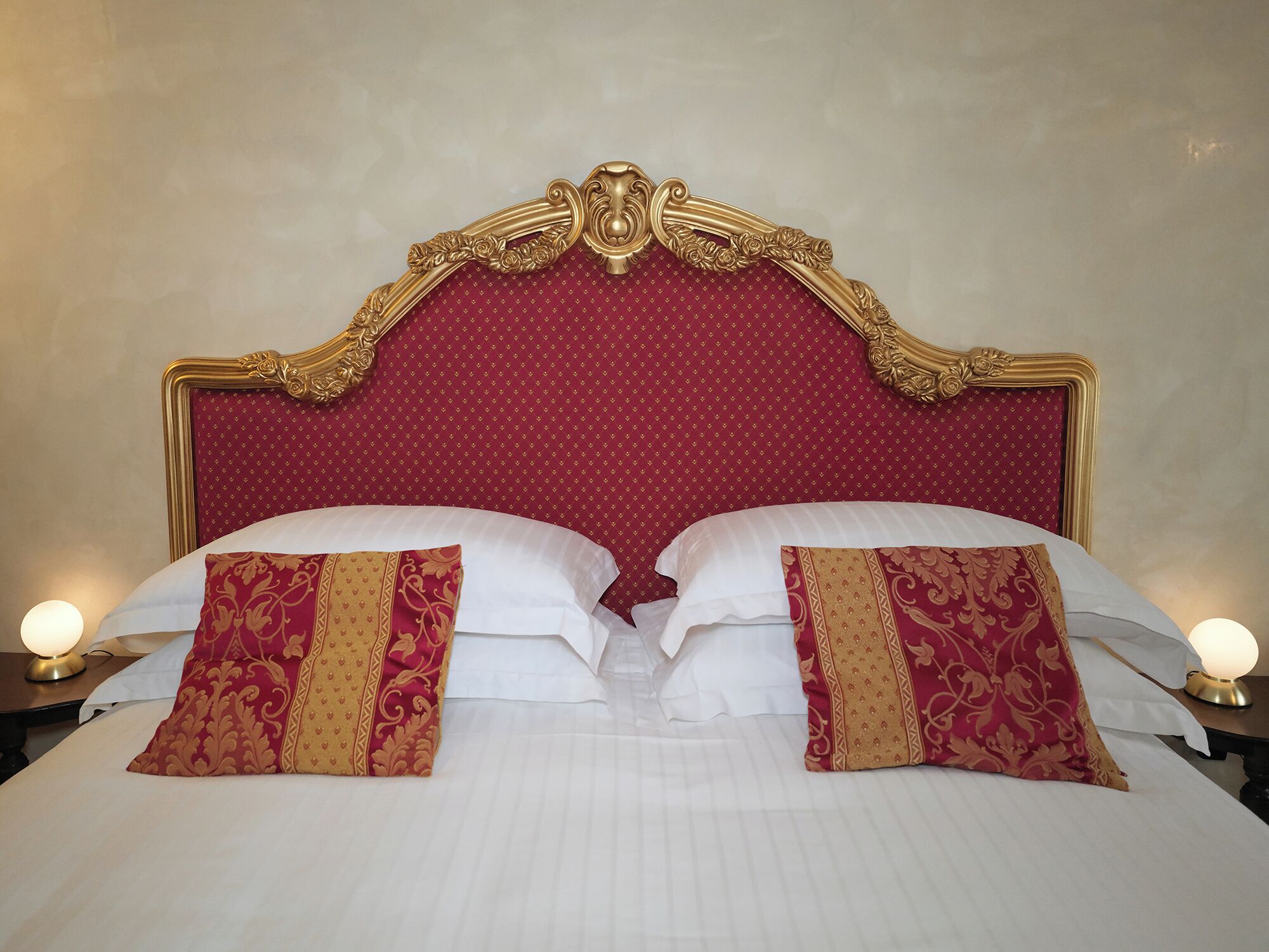 A bed with a red headboard and gold trim, two white pillows, and two decorative pillows with gold and red patterns.