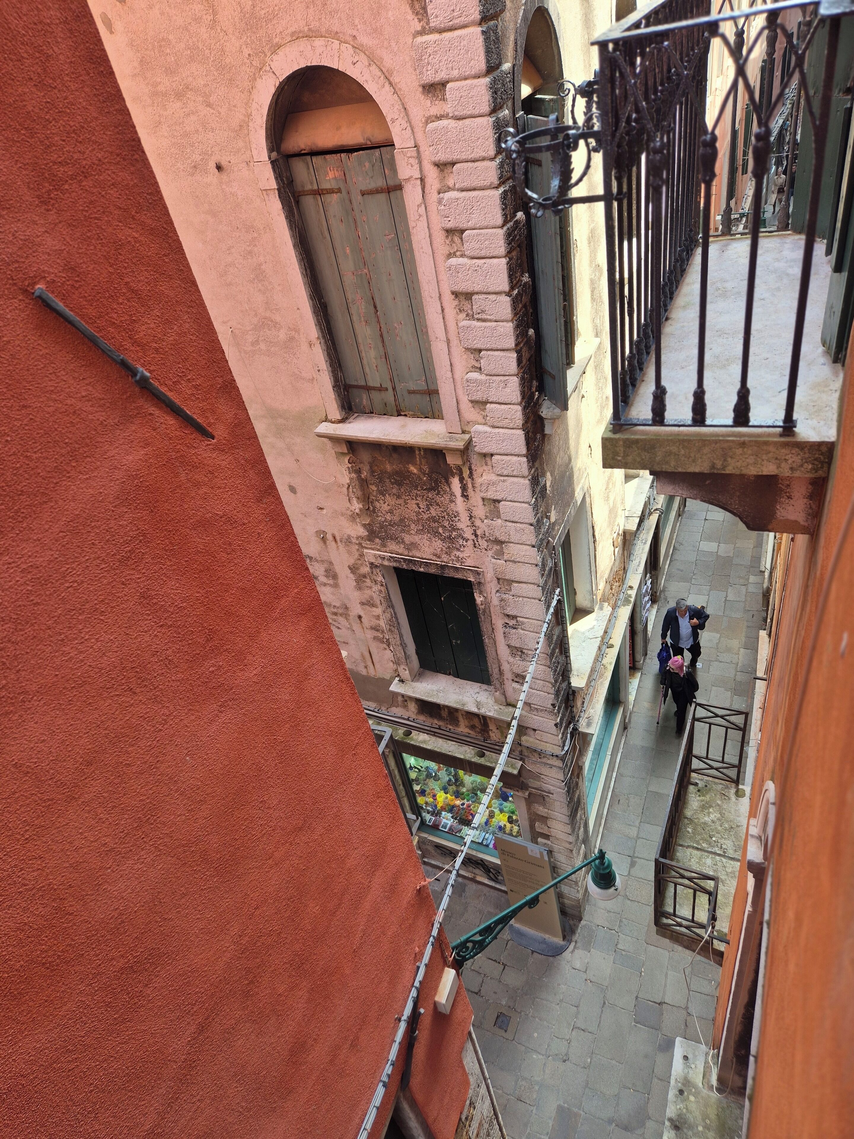 Une ruelle étroite avec un balcon, une fenêtre dotée d’un volet en bois, et une personne qui marche.