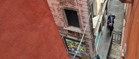 Une ruelle étroite avec un balcon, une fenêtre dotée d’un volet en bois, et une personne qui marche.