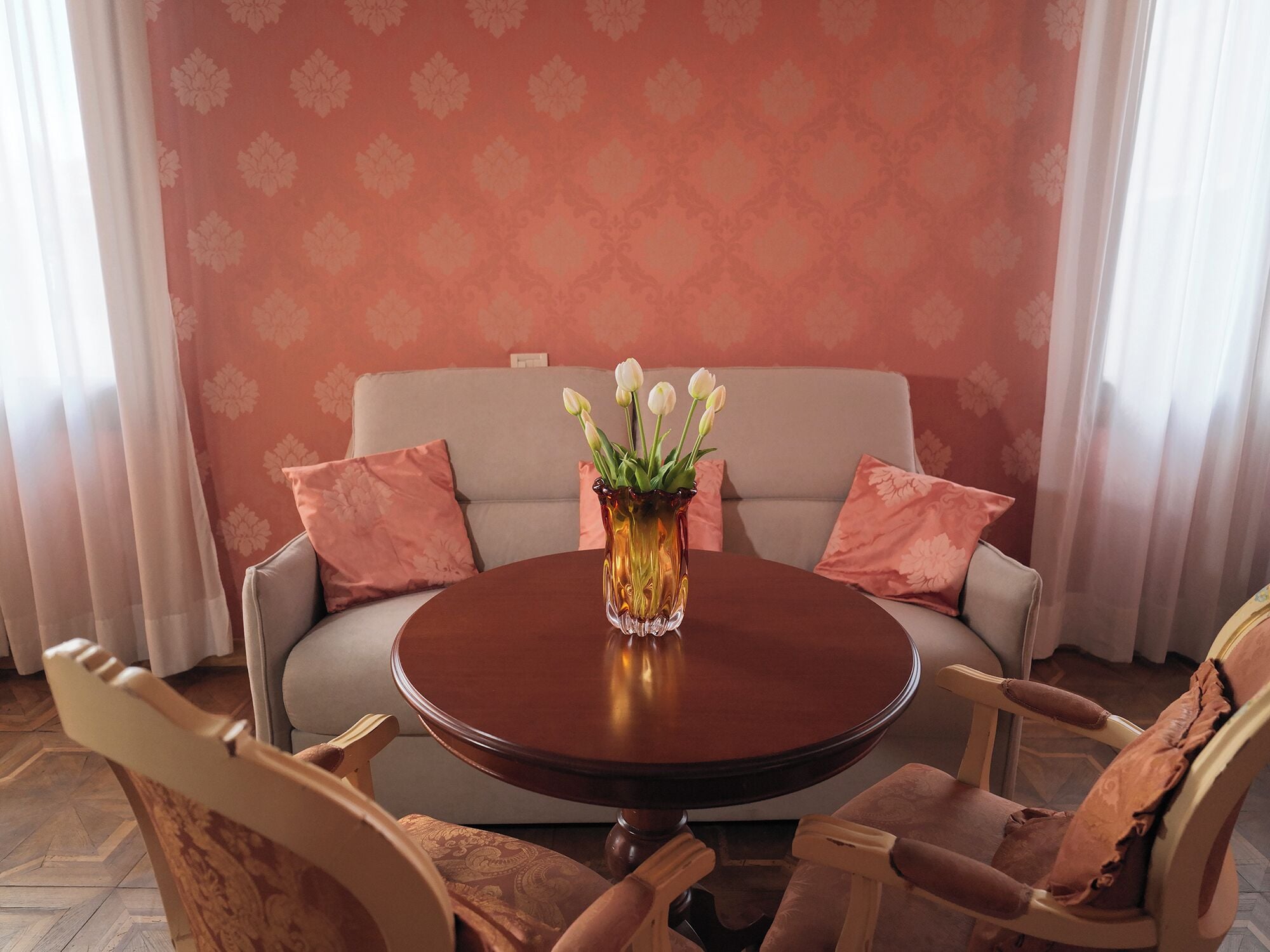 Una mesa redonda de madera con un jarrón de flores blancas, rodeada por cuatro sillas con tapicería estampada, frente a un muro decorado y un sofá con cojines a juego.
