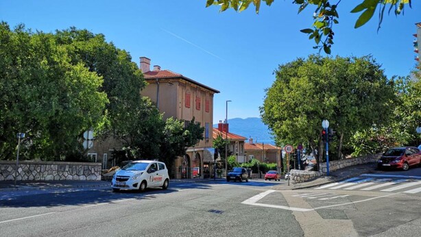 A street view with a white car parked on the side, a stone wall, and a building with a red roof in the background. - City Apartments Iva Krimeja Rijeka (Rijeka)