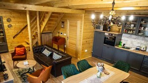 L’intérieur chaleureux d’un chalet en bois comprend un espace de vie avec un canapé et des fauteuils, une table à manger, une cuisine équipée d’appareils modernes et un escalier menant à l’étage supérieur.