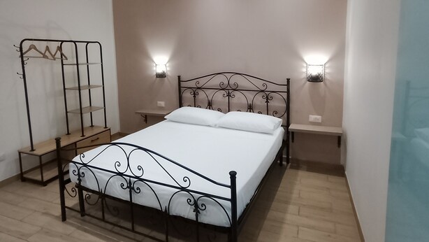 Un dormitorio con cama, mesitas de noche, luces fijadas en la pared y una estantería metálica.