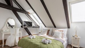A bedroom with a sloped ceiling, a bed with a green quilt, a TV mounted on the wall, a bedside table with a lamp, and a window providing natural light. - Zagreb Downtown Luxury Stay – 5th Element (Zagreb)