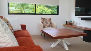 A modern living room with a sofa, a coffee table, a TV, and a window with a view of greenery. - Villa 3 suites, private pools, comfort & relaxation (Provincia de guanacaste)