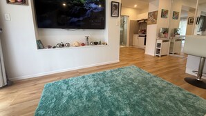 Ein modernes Wohnzimmer mit einem an der Wand montierten Flachbildfernseher, einem grünen Teppich und einem durch eine offene Tür einsehbaren Küchenbereich.