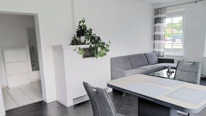 A modern living room with a grey sofa, a dining table with chairs, and a potted plant on a white cabinet. - Comfort vacation apartment Niemann 4 with private garden/terrace (Lübeck)